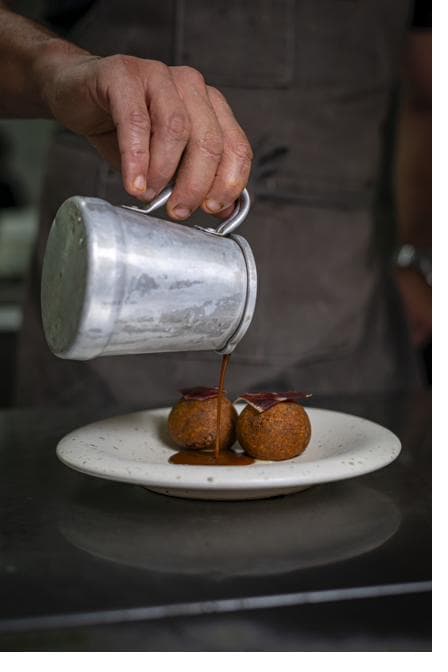 Chef a regar arroz de pato com molho, servido em tacho de ferro sobre tábua de madeira